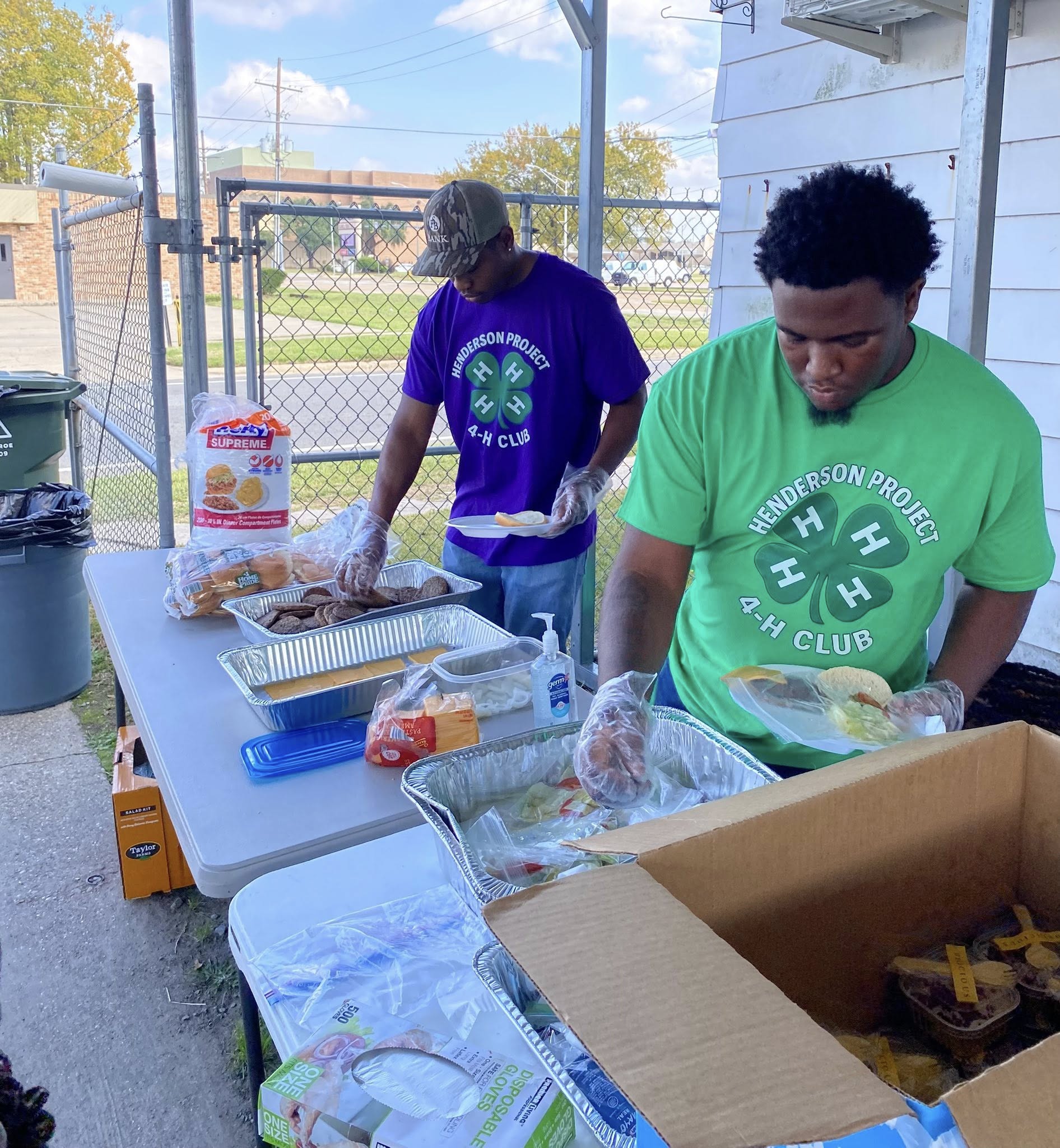 Two men wearing 4-H T-shirts putting food on plates. 4-H Emblem logo.